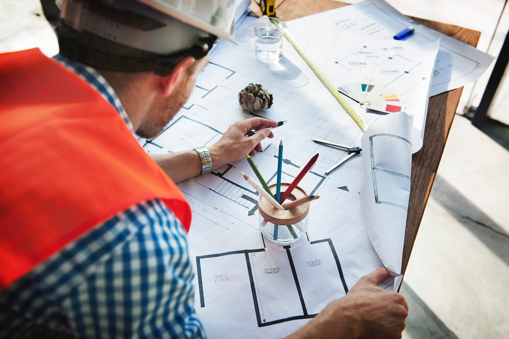 Antino and Associates employ expert party wall surveyors. Image of a surveyor with a high-vis jacket and hard hat leaning over some floor plans on a table.